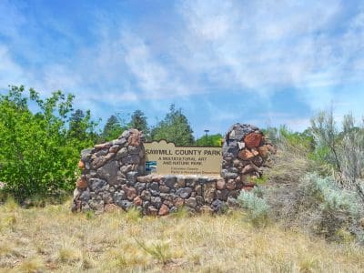A stone sign at the entrance of Sawmill County Park reads “A Multicultural Art and Nature Park” and is surrounded by grass, shrubs, and trees under a partly cloudy sky.