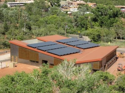A modern, single-story building with a red roof houses the Sedona-Oak Creek USD District Office PV System, featuring multiple rows of solar panels on top, surrounded by greenery and situated near a road.