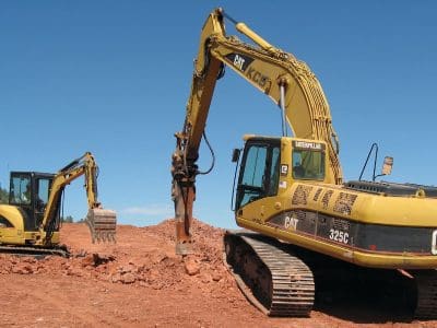 Two yellow excavators work on a construction site, moving earth on a clear, sunny day with a blue sky in the background.