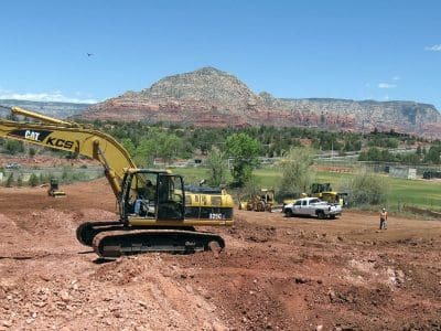 Yellow excavator digging in a dirt construction site with trucks and workers present; red rock mountains and a small field are visible in the background under a clear sky.