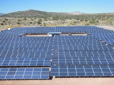 Rows of solar panels, part of the Sedona Wastewater Reclamation Plant PV System, are installed in an open field with hills and vegetation in the background on a clear sunny day.