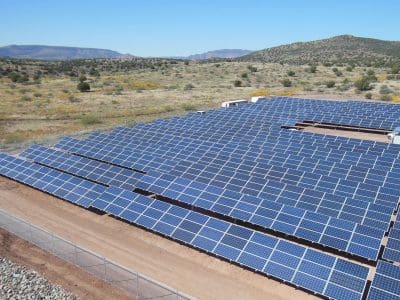 Rows of solar panels installed on a fenced plot of land in a rural area, part of the Sedona Wastewater Reclamation Plant PV System, surrounded by hills and sparse vegetation under a clear blue sky.