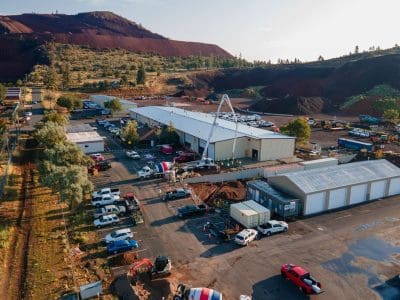 Aerial view of an industrial construction site with several buildings, vehicles, construction equipment, and a rocky hill in the background.