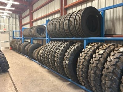 Rows of large industrial tires are stacked on blue metal racks inside a warehouse with concrete floors and corrugated metal walls.