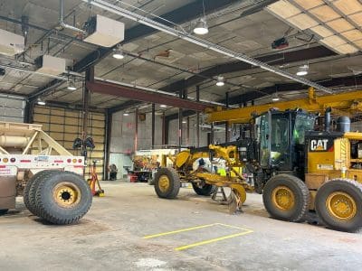 A large industrial workshop with heavy equipment, including a CAT grader and a trailer, surrounded by tools and machinery under overhead lights.
