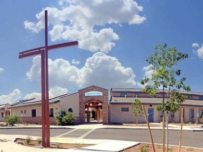 A modern one-story church building with a large red cross in front, surrounded by a paved parking area, under a bright blue sky with scattered clouds.