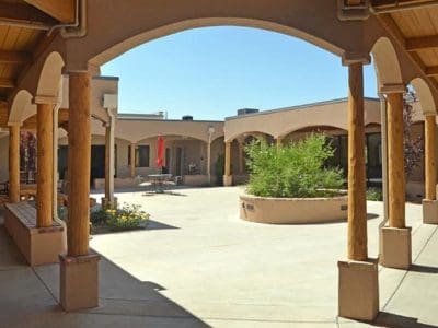 A view of a shaded courtyard with adobe-style architecture, wooden columns, arched walkways, and a central planter with greenery.