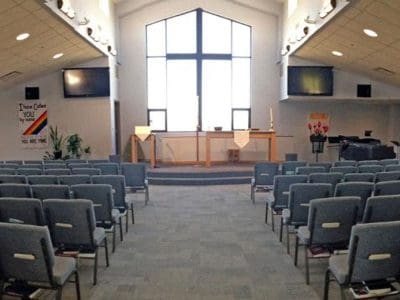 Interior of a modern church with rows of gray chairs facing an altar, large cross-shaped window, banners on each wall, and screens mounted on both sides.