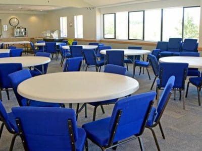 A room with multiple round tables and blue chairs arranged around them, featuring large windows and a refreshment area in the background.