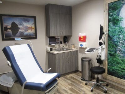 A clean, modern medical exam room with an exam table, sink, cabinets, medical supplies, computer station, and nature-themed wall art.