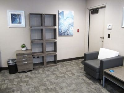 A modern office corner with an empty wooden shelf, a small cabinet with a plant, a gray armchair, a side table, wall art, and a closed door.