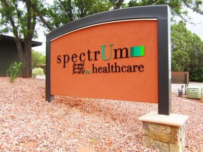 A large outdoor sign reads "spectrum healthcare" with the tagline "for mind, for body, for all of you" on a textured brown background, surrounded by rocks and trees.