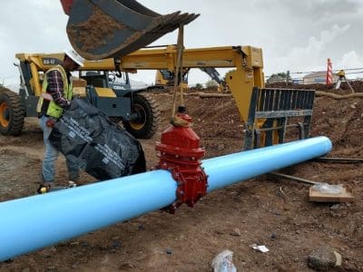 A construction site with workers installing a large blue water pipe using heavy machinery; a red valve is attached to the pipe.