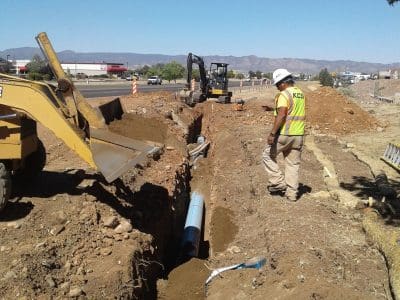 A construction worker observes an excavator and bulldozer digging a trench and laying a blue water pipe at a roadside construction site.