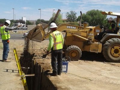 Three construction workers in safety gear operate a backhoe loader to pour dirt into a trench at a roadside construction site.