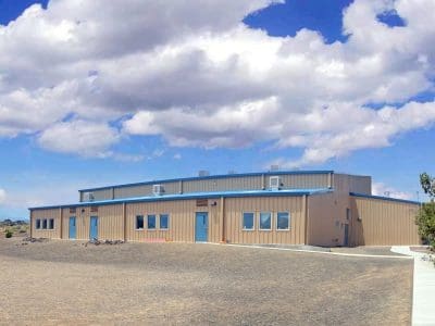 A tan and blue industrial building sits on a barren lot under a partly cloudy sky, with sparse vegetation and a paved walkway leading to the entrance.