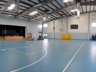 A large indoor gymnasium with blue flooring, basketball hoops, bleachers, a stage with flags, and stacked equipment near the stage.