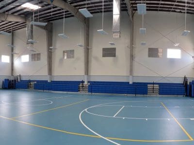 Empty indoor gymnasium with blue bleachers, basketball hoop, and marked court lines on a light blue floor.