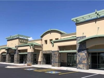 A modern, empty strip mall with multiple storefronts, stone accents, and green trim, viewed from a parking lot with handicap spaces under a clear blue sky.