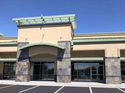A vacant commercial building with large glass windows, stone accents, and green metal trim, set against a clear blue sky.