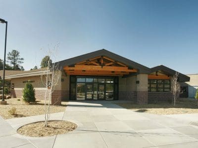 Single-story modern building with large windows, brick and wood exterior, paved walkway, and minimal landscaping under a clear sky.