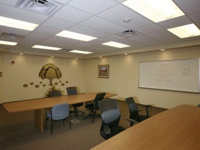 A conference room with two long tables, several office chairs, a whiteboard with writing, wall decorations, and overhead fluorescent lights.