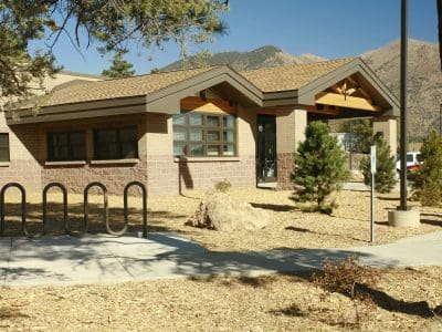 Single-story building with large windows and a bike rack in front, surrounded by trees, in a dry, mountainous landscape under clear blue sky.