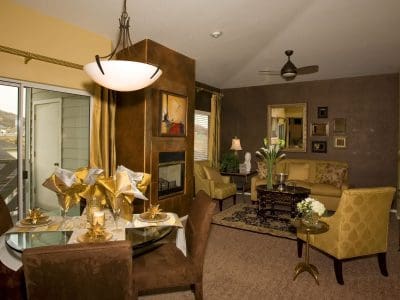 A modern living and dining area features a round glass table with four chairs, a fireplace, contemporary lighting, and gold-toned decor, with large windows and a balcony view.
