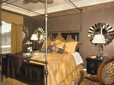A bedroom with a four-poster bed, gold and brown bedding, two sunburst mirrors above nightstands with lamps, a leopard-print chair, and a window with gold curtains.