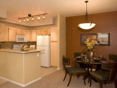 A small modern kitchen with light wood cabinets and a white refrigerator is adjacent to a dining area with a round table set for four against a brown accent wall.
