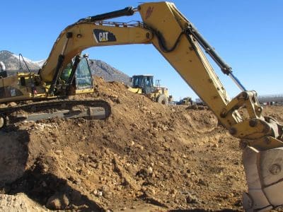 A yellow excavator moves dirt at a construction site, with a bulldozer and mountains visible in the background under a clear blue sky.