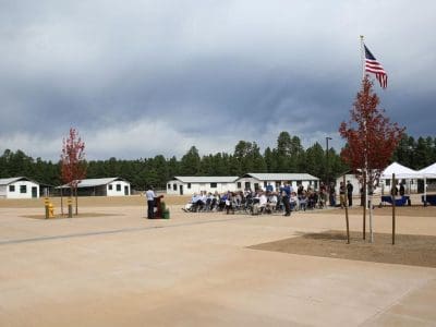 A small group of people sit in folding chairs near white buildings, while an American flag flies in the foreground on a cloudy day.