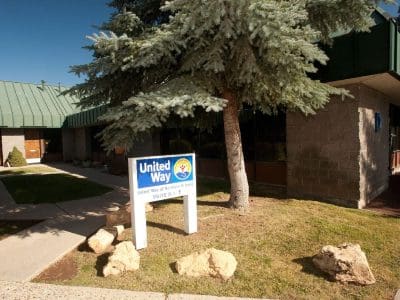 A United Way sign stands outside a single-story building with a green roof, surrounded by trees and landscaping on a sunny day.