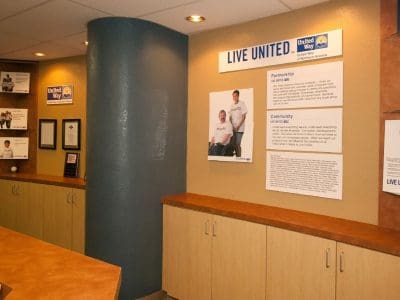 Reception area with beige counters and cabinets, United Way "LIVE UNITED" signs and posters on tan walls, informational brochures, and framed certificates on display.