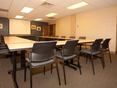 A conference room with a U-shaped table arrangement, several black chairs, a projector screen, framed pictures on the wall, and a potted plant in the corner.