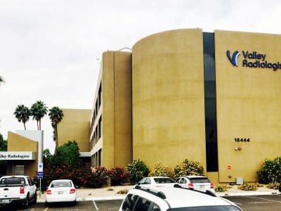 A beige medical building labeled "Valley Radiologists" with parked cars and palm trees in front.