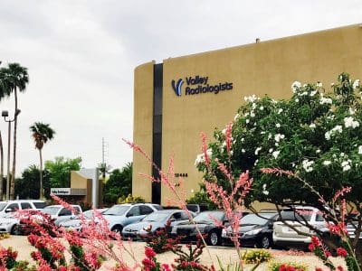 A tan building labeled "Valley Radiologists" stands behind a parking lot with several cars and flowering trees in the foreground.
