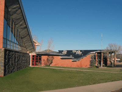 A modern brick building with large windows and slanted roofs sits on a lawn under a clear blue sky.
