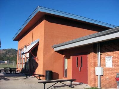A red brick and orange building with large windows, red double doors, outdoor picnic tables, and a metal awning, set against a clear blue sky.