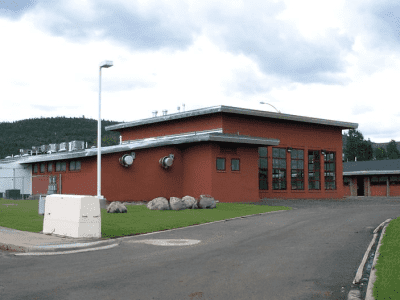A single-story industrial brick building with large windows and ventilation units sits on a paved road surrounded by grass, rocks, and distant trees under a cloudy sky.