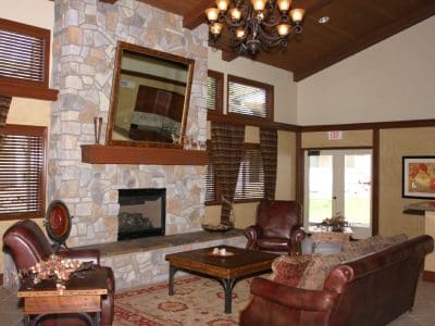 A living room with stone fireplace, two leather chairs, a sofa, a chandelier, rug, and large windows with drapes; kitchen area visible in the background.