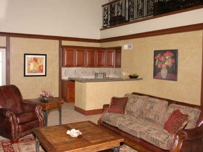 A living room with a leather armchair, patterned sofa, wooden coffee table, and a small open kitchen in the background. Artwork and a staircase railing decorate the walls.