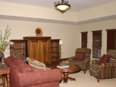 A living room with a red-brown sofa, two patterned armchairs, a wooden coffee table, a wooden entertainment center, and decorative lamps and plants.