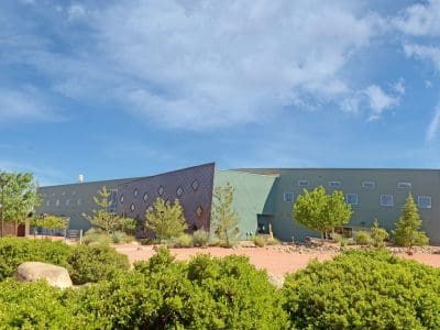 Modern building with geometric design elements, surrounded by desert landscaping with shrubs, rocks, and trees under a partly cloudy sky.