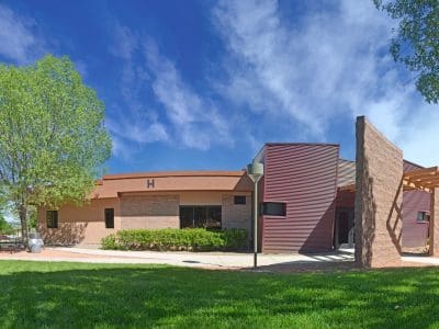 Single-story building with modern architectural features, surrounded by green grass, trees, and a clear blue sky. The letter "H" is visible above an entrance.