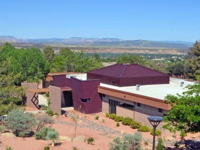 A modern, flat-roofed building with maroon accents is surrounded by desert landscaping and trees, with distant mountains visible under a clear blue sky.