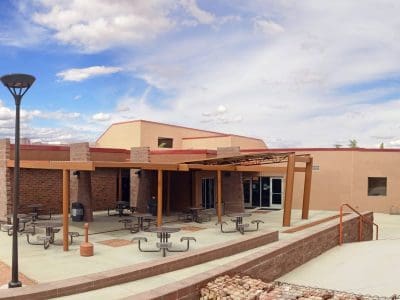 Single-story building with a shaded patio area, outdoor tables and benches, lamp post, and a partly cloudy sky in the background.