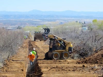 Construction workers operate heavy machinery and dig a trench along a dirt path surrounded by brush, with mountains visible in the background.