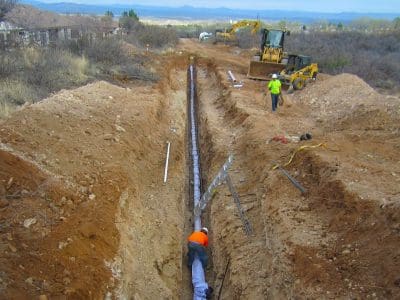 Construction workers are installing a pipeline in a deep trench, with excavators and equipment visible on the site surrounded by dirt and brush.