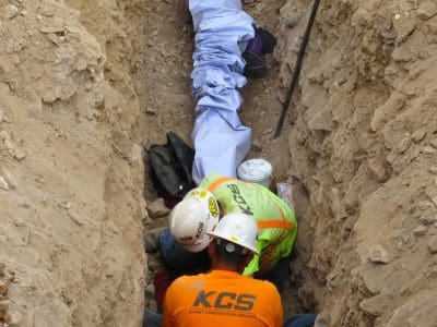 Two construction workers in safety gear attend to a person covered with a white sheet in a narrow dirt trench.